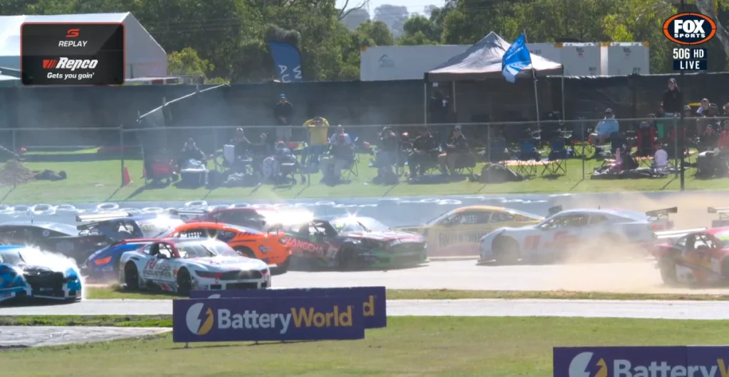 Trans-Am-Race-3-Crash-Fox- Sports • Sports Rewind Media Multiple race cars start a race on a sunny track, dust and smoke rising as they jostle for position while spectators sit on the grass behind a fence with tents.