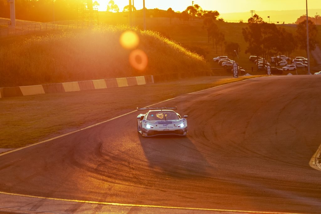 _I1A1685 • Sports Rewind Media White race car on a sunlit race track during sunset, headlights on and orange glare filling the scene.
