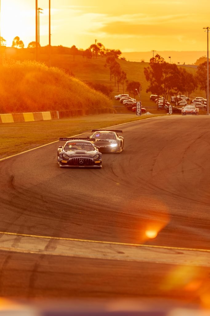 _I1A1720 • Sports Rewind Media Two race cars lead a curved track, bathed in orange sunset light with a distant pit area and hillside in the background.