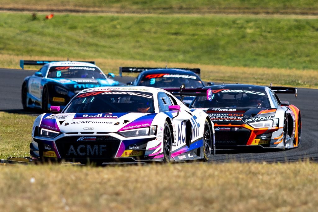 _I1A2384 • Sports Rewind Media Three colorful race cars racing in a tight pack on a sunny track with grass in the outfield, visible sponsor logos on the cars.