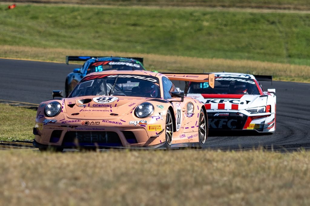 _I1A2401 • Sports Rewind Media Pink Porsche race car leads a pack on a curving track with green grass in the background, sponsor logos visible on the body.