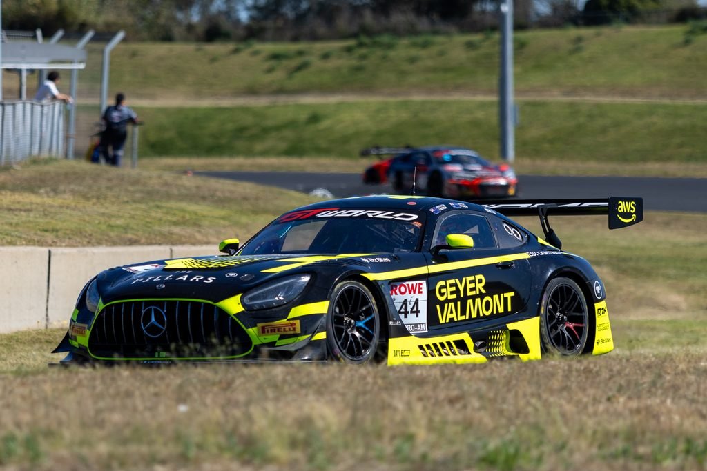 _I1A2410 • Sports Rewind Media Black-and-yellow race car in the foreground on a race track, with a second car and spectators behind a fence in the distance