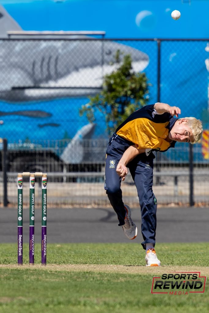 _I1A4603 • Sports Rewind Media Cricketer in yellow and navy delivering a ball toward the stumps on a sunny cricket pitch; ball in midair with a blue fence backdrop.