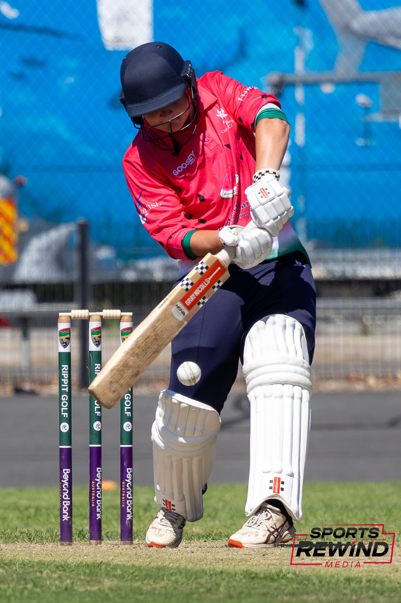 _I1A4662 • Sports Rewind Media Cricketer in a red shirt wearing helmet and pads, swinging a bat near the stumps during a shot at the crease.