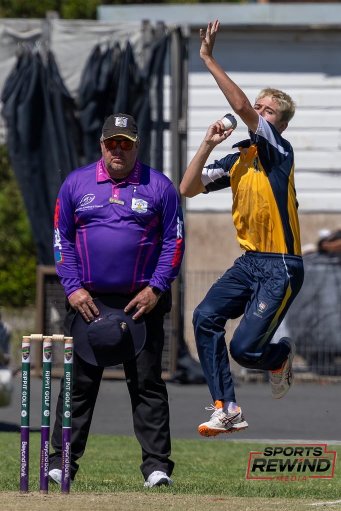_I1A4698 • Sports Rewind Media Cricket scene: umpire in purple shirt and cap watches as a bowler in yellow and navy leaps to deliver the ball on a sunny field, with stumps in the foreground and a blurred backdrop.