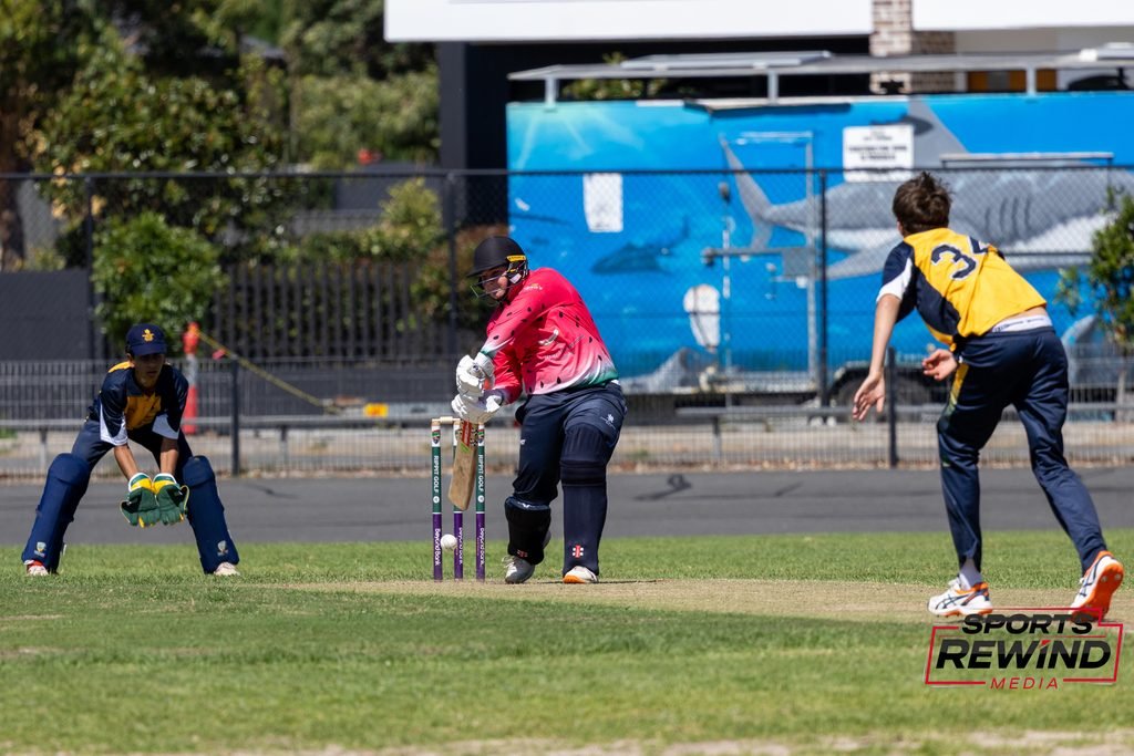 _I1A4770 • Sports Rewind Media Cricketer in a red shirt at the crease prepares to bat as two fielders in navy and yellow guard the field, with a blue fence in the background and a 'Sports Rewind' logo bottom right.