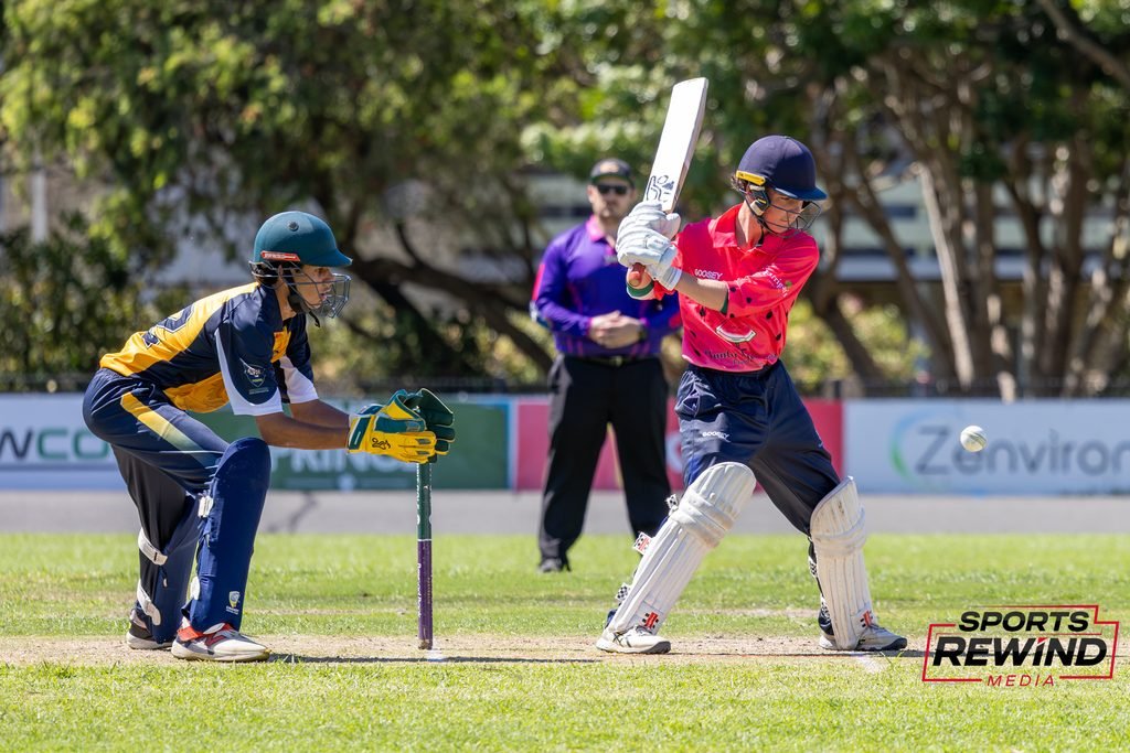 _I1A4781 • Sports Rewind Media Cricketers in action: a batsman in pink swings at the ball while a fielder/keeper in blue and yellow crouches behind him on a sunny field with a referee in the background.
