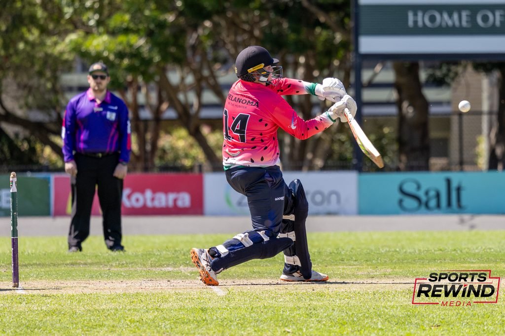 _I1A4801 • Sports Rewind Media Cricket batsman in a pink jersey swings at the ball during a match, wearing a helmet and pads with an umpire watching in the background.