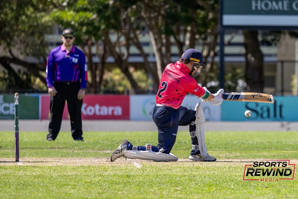 _I1A4809 • Sports Rewind Media Cricket batsman in red kneels at the crease, helmeted and padded, swinging a bat with an umpire in a purple shirt in the background.