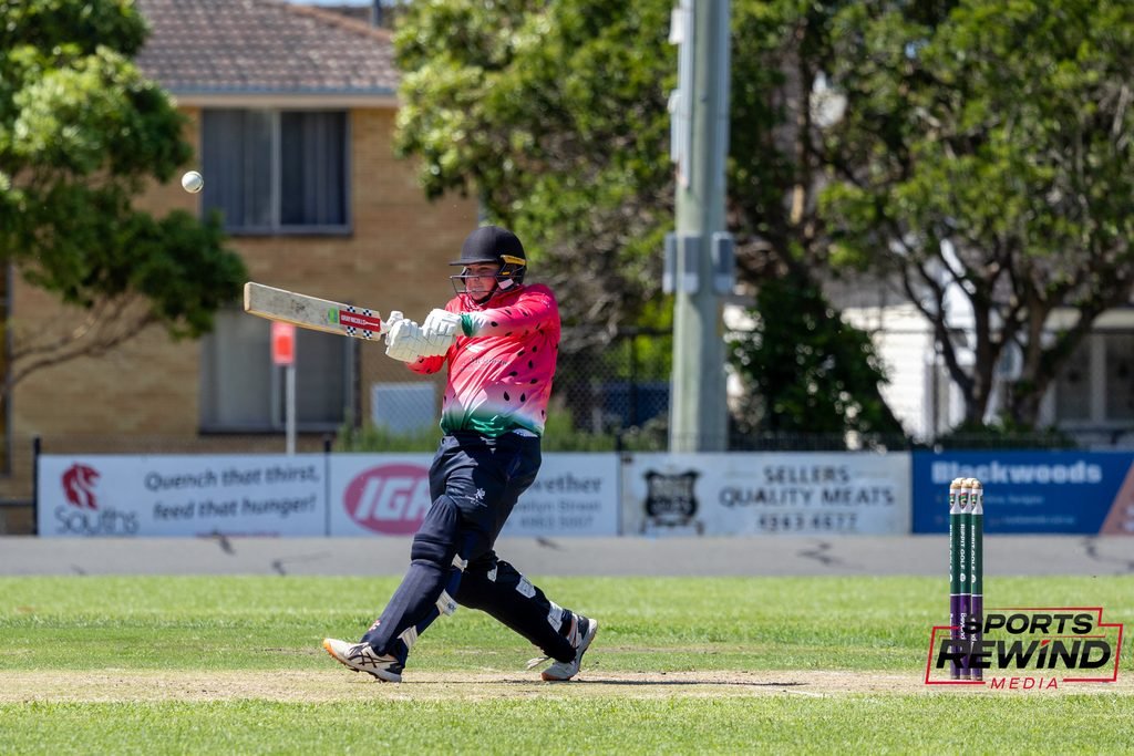 _I1A4838 • Sports Rewind Media Cricketer in a pink jersey and helmet swings a bat at fast-moving ball on a sunny field, wickets in the foreground.