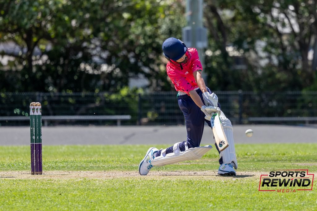 _I1A4871 • Sports Rewind Media Cricketer in a pink jersey batting, helmeted, on a sunny field with stumps nearby.